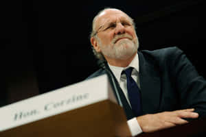 一位身穿西裝、留鬍鬚並戴眼鏡的男子，在標有 Hon Corzine 的講台上向上看。 — 政治 壁紙
