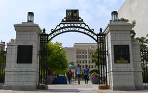 人們在陽光明媚的日子裡穿過寫有 Professors Gate 的大學大門。