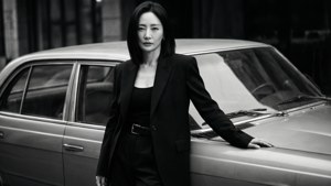 一位身穿黑色西裝的女士在黑白照片中靠在一輛復古車旁。