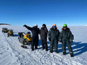 四個穿著冬季裝備的人在雪地上與雪地摩托車合影。 — 日出 壁紙