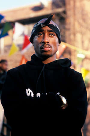 Tupac Shakur 身穿黑色連帽衫和頭巾，戶外站立，身後有色彩繽紛的旗幟。