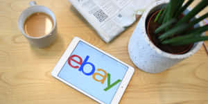 平板電腦顯示eBay標誌在木桌上，附有咖啡杯、報紙和植物。