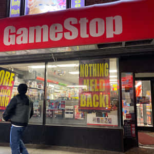夜晚，一個人走過一間有明亮招牌和海報的GameStop商店。 — 遊戲 壁紙