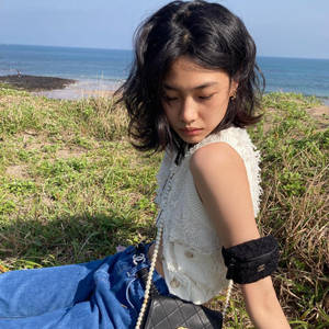 身穿白色上衣和藍色牛仔褲的年輕女性，日落時分坐在海邊草地上。 — 韓國 壁紙