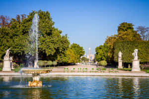 陽光明媚的公園中的大型噴泉，背景有雕像和城市天際線。