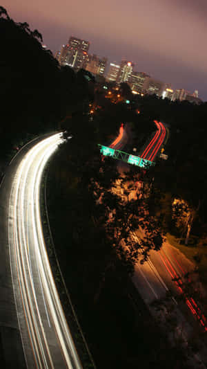 夜間高速公路，移動的汽車在靠近城市建築的樹木間蜿蜒行駛，留下光跡。