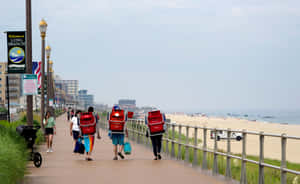 三名背著紅色背包的送餐員在海灘附近的步道上行走。