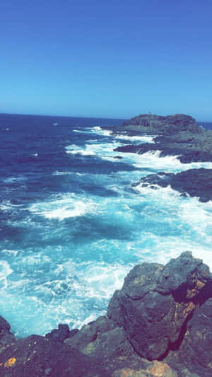 在晴朗的藍天下，藍綠色的海浪拍打在深色火山岩上。