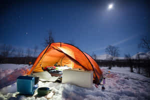 在繁星點點的夜空下，雪地上有一頂溫暖照亮的橙色帳篷和一輪明亮的月亮。 — 露營 壁紙