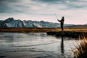 漁夫在河邊拋出魚線，背景有白雪覆蓋的山脈