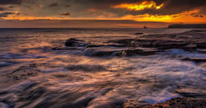 夕陽映照海浪拍打岩石海岸，遠方有燈塔