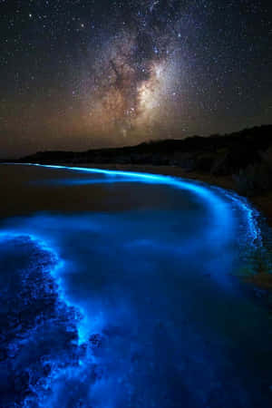 在銀河下的星空下，黑暗沙灘上生物螢光波浪發出藍光。 — Vivo Pad SE 壁紙