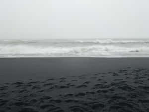 在灰色天空下，暴風雨的海灘上，黑色火山沙中的腳印，海浪拍打著。 — 海灘日落 壁紙