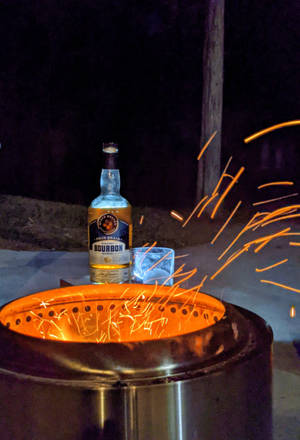 夜間營火旁的波本威士忌瓶和玻璃杯，火花飛舞。 — 夜晚 GIF 壁紙