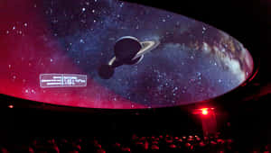 觀眾在彎曲的圓頂屏幕上觀看以土星和宇宙數據為特色的天文館表演。 — Astroworld GIF 壁紙