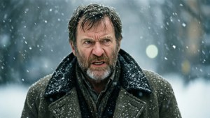一個留鬍子的男人穿著冬季外套，站在雪景城市場景中，表情嚴肅。