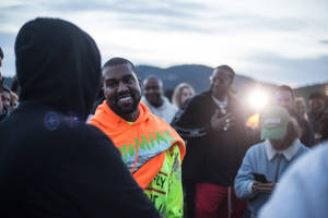 Kanye West 喺戶外活動中，身穿鮮艷連帽衫微笑，背後有山。