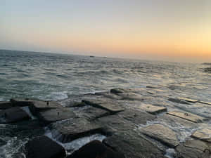 海浪拍打石堤，夕陽下遠方船隻在平靜海平線上