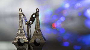 兩個刻有「Paris」的小型艾菲爾鐵塔鑰匙扣，掛在模糊的藍色燈光背景上。 — 巴黎鐵塔 壁紙