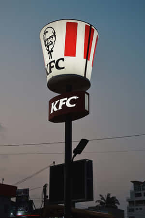 KFC桶形招牌在黃昏時分於城市環境中亮起，伴隨棕櫚樹和建築物。 — 小米Pad 7S Pro 12.5 壁紙