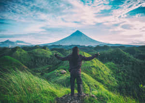 雙臂張開的人站在山丘上，俯瞰綠色山谷和遠方的火山。