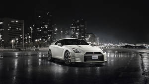 白色日產GT-R夜間停在濕漉漉的城市街道上，背後是發光的建築物。