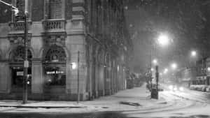 夜間城市街道上飄雪，有明亮的店面和停泊的汽車。