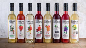 八個帶有插圖標籤的 Urban Cordial 水果味飲料瓶，放在木桌上。 — 精細 壁紙