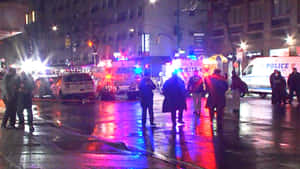 緊急應變人員和旁觀者在夜間雨中的城市街道上，伴隨著閃爍的燈光。