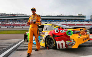 身穿橙色M&M's服的NASCAR車手站在賽車道上，旁邊是他色彩繽紛的賽車。 — 賽車 壁紙