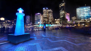 夜晚城市廣場上的藍色發光燈塔雕塑，背後是燈火通明的摩天大樓。