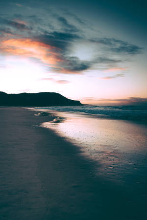 夕陽映照在平靜的海灘上，有海浪和剪影般的山丘 — 海灘日落 壁紙
