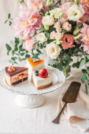 三件甜品切片放在白色蛋糕架上，附近有一束花。