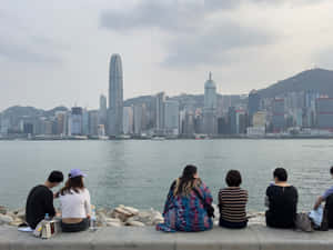 五個人坐在石壆上，在陰天觀看香港的天際線和港口。 — 天際線 壁紙