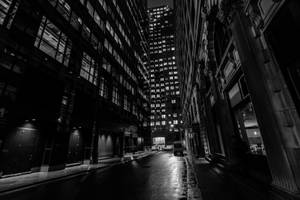 夜晚黑暗的城市小巷，高樓大廈和發光的窗戶在濕漉漉的路面上。 — 黑白 壁紙
