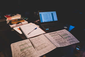 夜晚昏暗燈光下，雜亂的書桌上放著打開的筆記本、筆記本電腦和書籍。 — MacBook Pro M2 壁紙