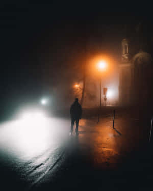 一個人在霧氣瀰漫的夜晚街道上獨自行走，被路燈和車頭燈照亮。