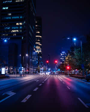夜晚城市街道，發光的建築物和紅色交通燈在潮濕路面上反射。