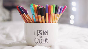 白色馬克杯上寫著「I DREAM IN COLOURS」，裝著彩色記號筆，放在床上。
