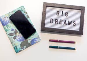 手機放在花卉圖案手機殼上，旁邊有寫著 big dreams 的字板和兩支筆。 — 勵志桌面 壁紙