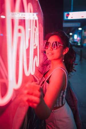 戴太陽眼鏡的女士在夜晚的城市街道上，在發光的紅色霓虹燈牌旁邊微笑。 — 霓虹美學 壁紙
