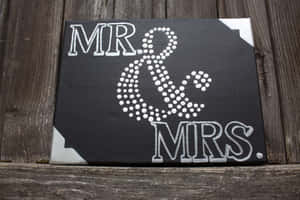 黑色畫布上印有白色 'MR & MRS' 文字及圓點和號，木質表面。