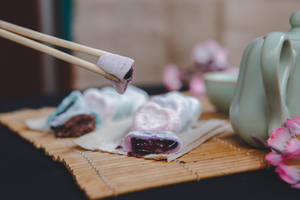 筷子撿起一個帶深色餡料的紫色麻糬糰子，旁邊有綠色茶壺和粉紅櫻花。 — 心形 壁紙