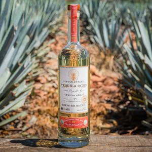 一瓶Tequila Ocho Añejo放在木質表面上，後面有龍舌蘭植物。