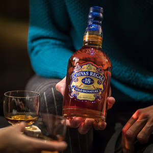 人拿著Chivas Regal威士忌酒瓶，旁邊放著一杯威士忌。