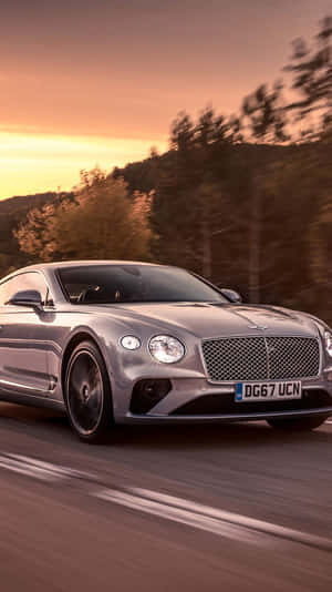 銀色賓利 Continental GT 在日落時分在道路上疾馳，背景有模糊的樹木。