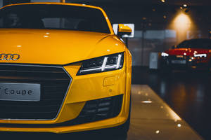 近距離拍攝一輛黃色Audi T Coupe，位於光線昏暗的汽車展廳，背景中有一輛紅色汽車。