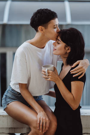 兩位女性擁抱，一人親吻另一人的額頭，同時手持杯子。