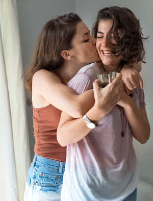 兩位女士擁抱大笑，其中一位拿著咖啡杯親吻另一位臉頰。