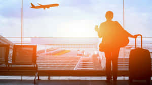 旅客帶著行李箱在夕陽下透過機場窗戶觀看飛機起飛。